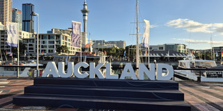 Auckland Ein großes Schild mit der Aufschrift „Auckland“, das sich im Hafen der Stadt befindet und hinter dem die Skyline zu erkennen ist.